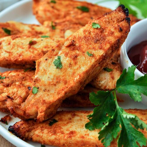 A close up of a stack of chicken tenders served with ketchup and a sprig of parsley.