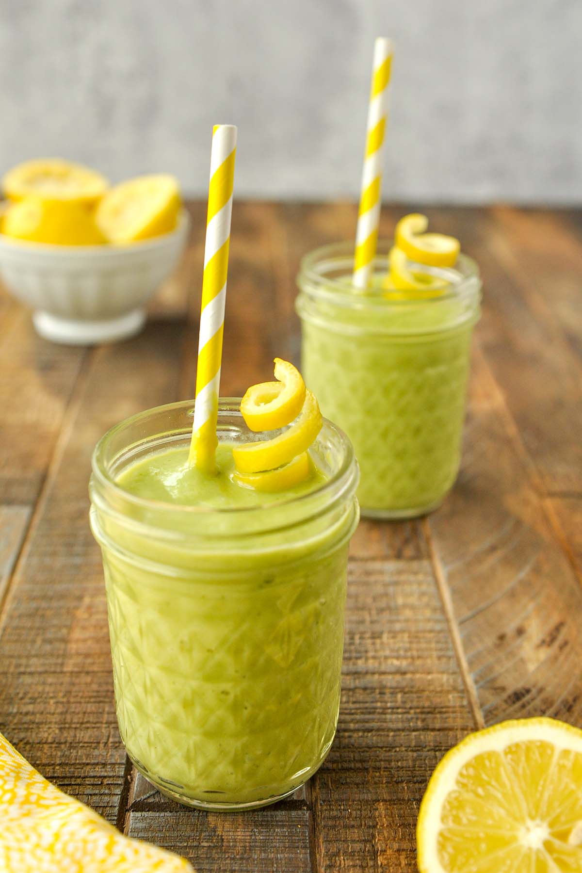 Two cups of lemon green smoothie with a twist of lemon peel and a yellow and white striped straw.