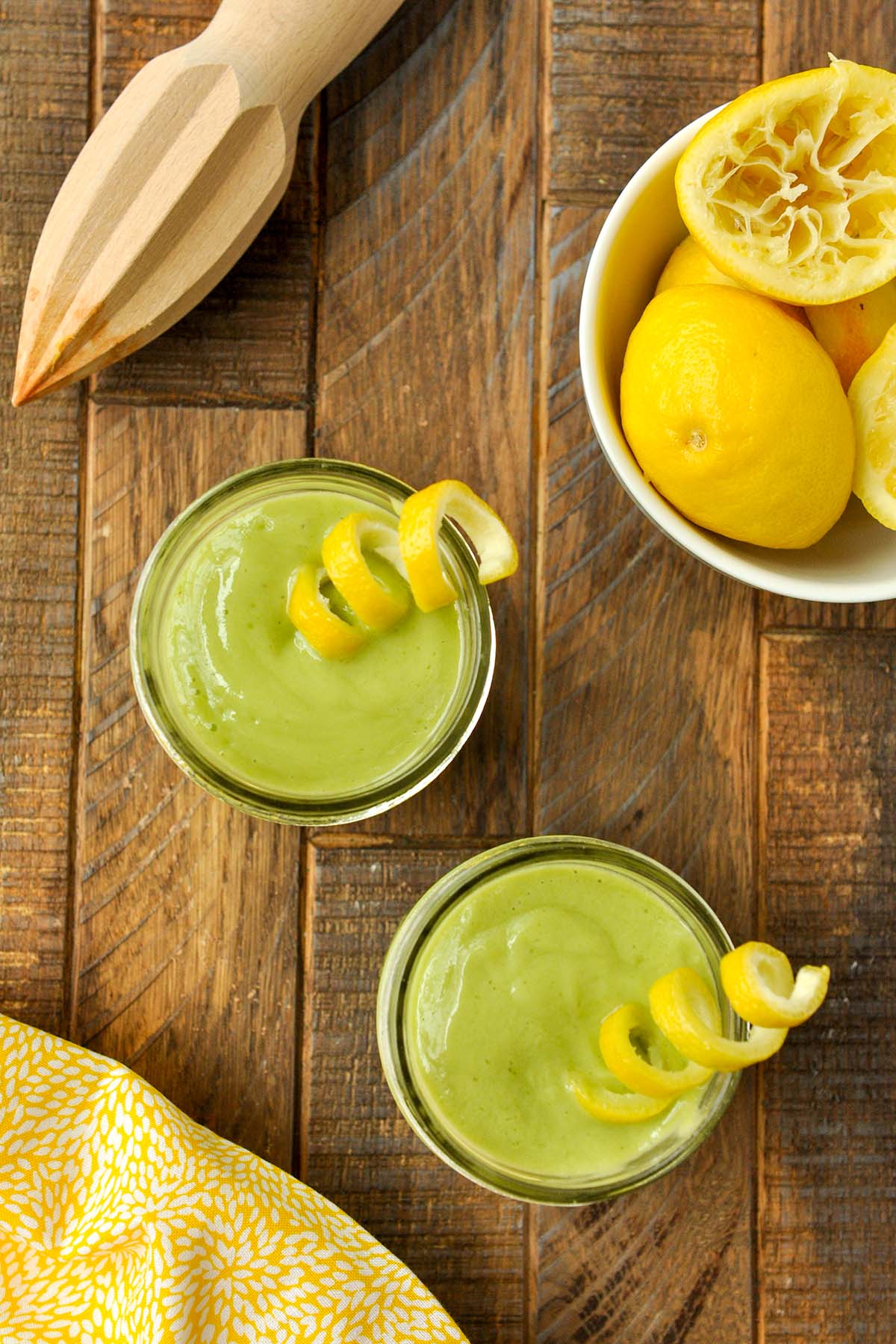 Above view of two cups of lemon green smoothie with a twist of lemon peel, a bowl of spent lemons and a lemon reeder.