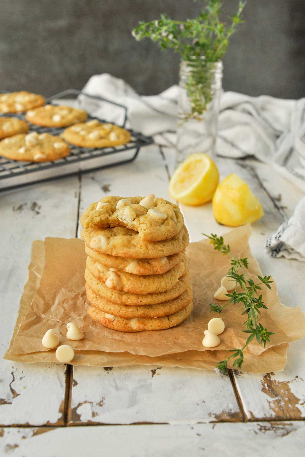 A stack of 7 cookies on a piece of parchment with white chocolate chips. Lemons and tray of cookies in the background.