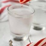 Two shot glasses of the polar bear shot with a peppermint stick above one glass with a drip coming down.