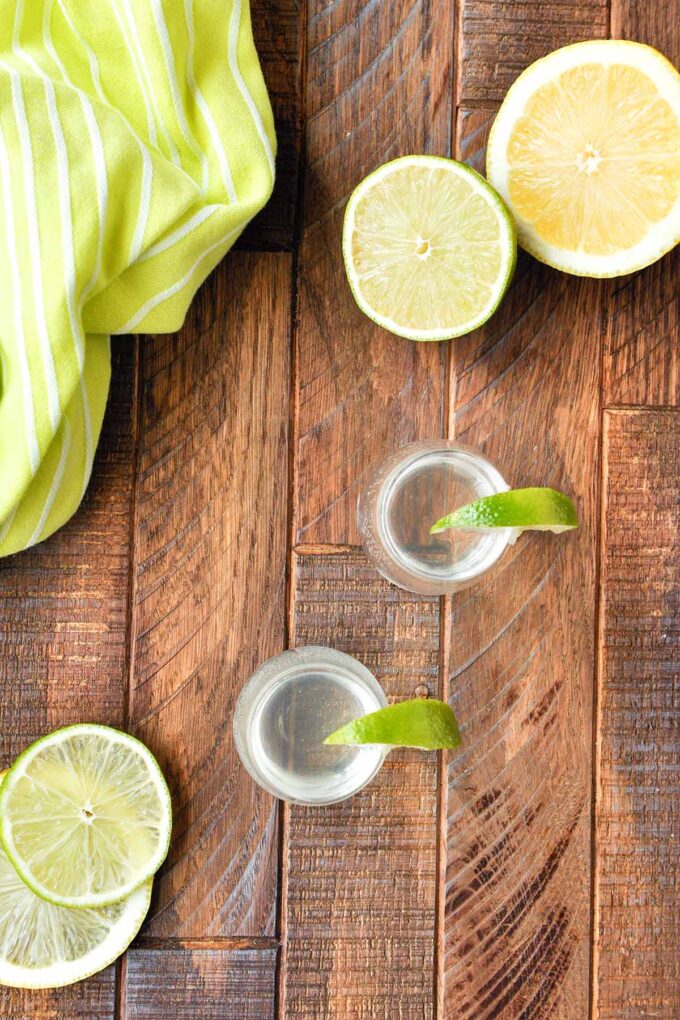 Above view of two shot glass with white tea shots and lime wedges, a green striped towel, a lemon half and lime half.