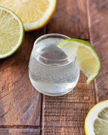 A single shot glass of the white tea shot with a lime wedge on the side of the glass and lemon and lime halves in the background.