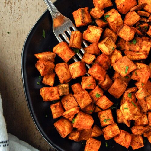 Above view of a plate of air fryer sweet potato cubes sprinkles with chopped parsley with a fork and a towel.