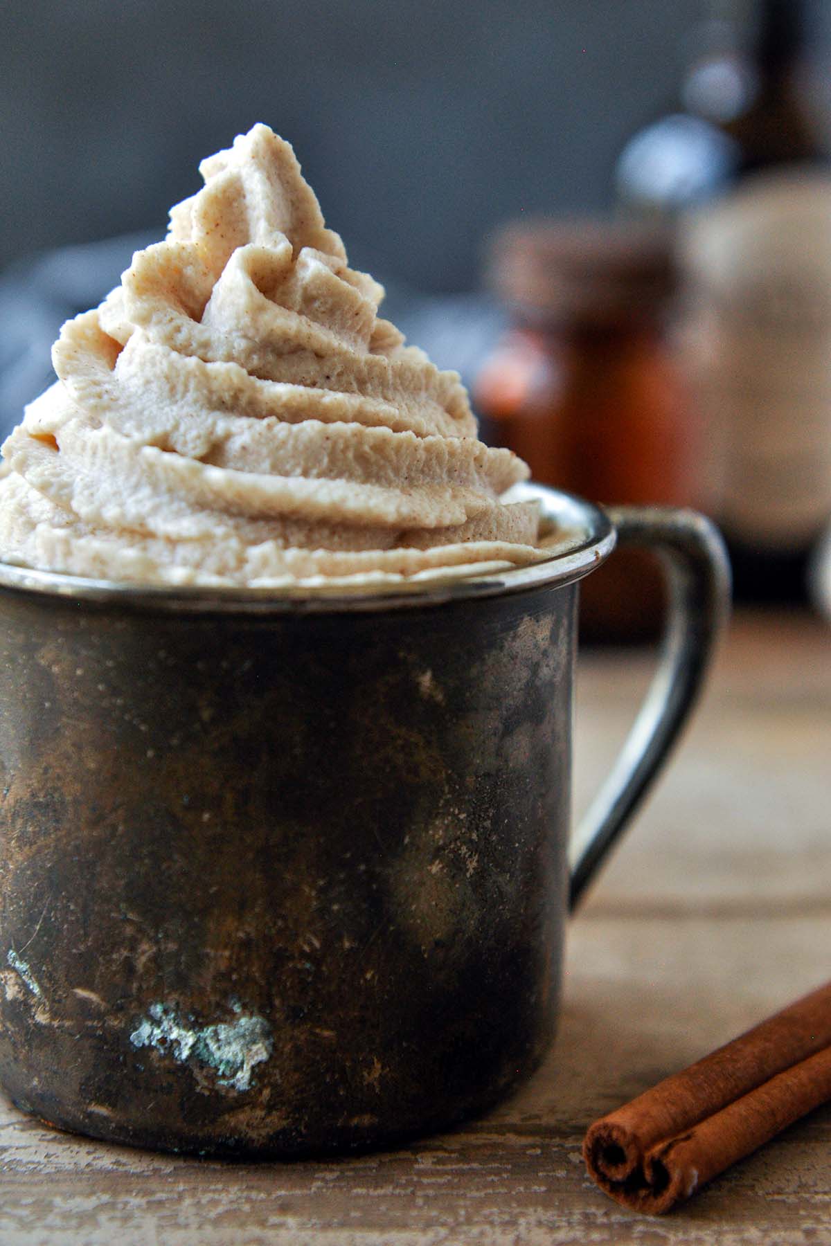 A very up close view of a metal cup filled with a swirl of cinnamon whipped cream, glass bottles in the background with a cinnamon stick up front.