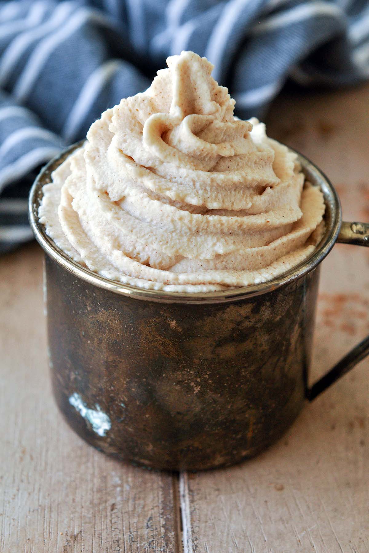 A metal cup filled with a swirl of cinnamon whipped cream with a black striped towel.