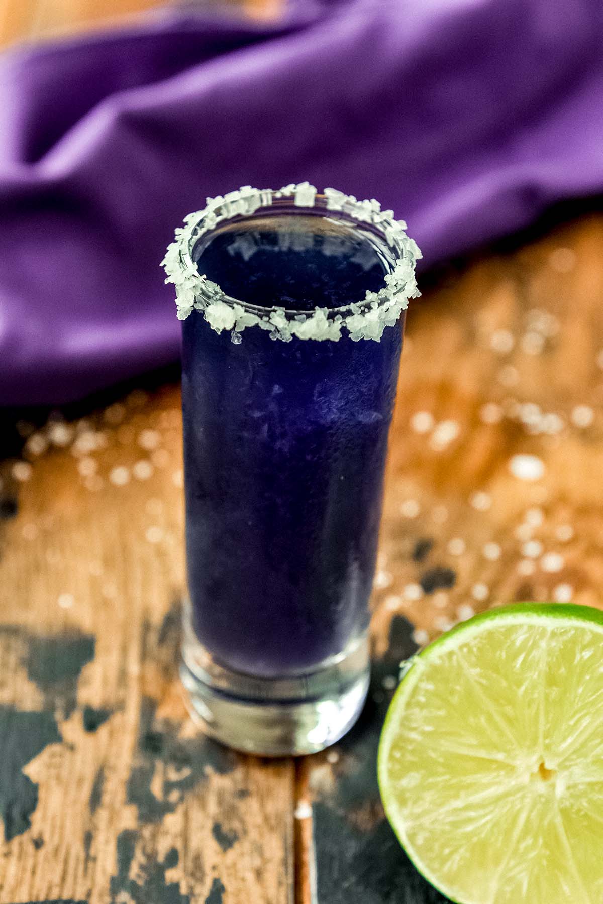 An up close of a shot glass with purple cocktail and a lime half.
