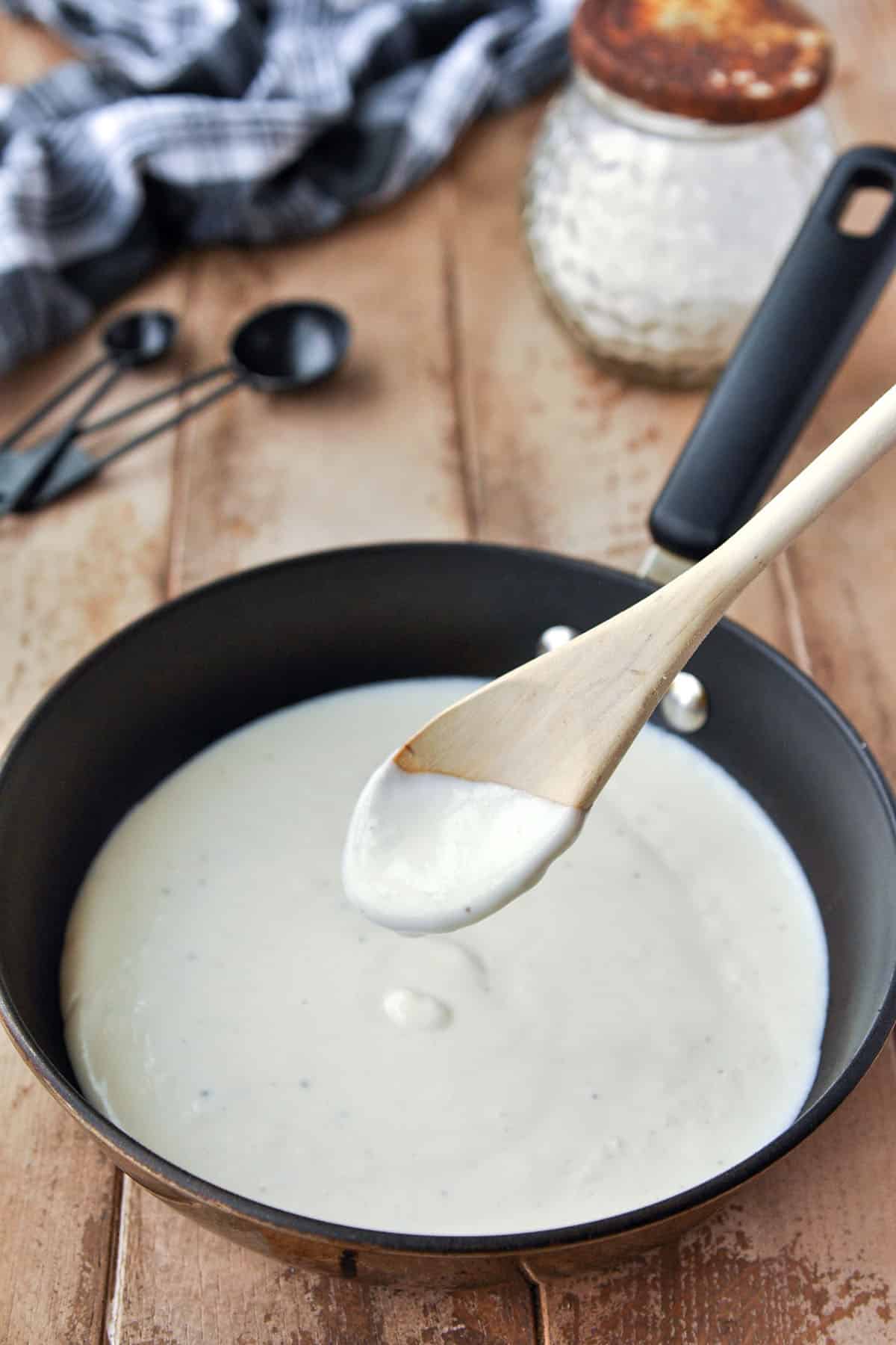 A pan of salsa bechamel with a spoonful of sauce. A towel, jar of flour, and measuring spoons in back ground.