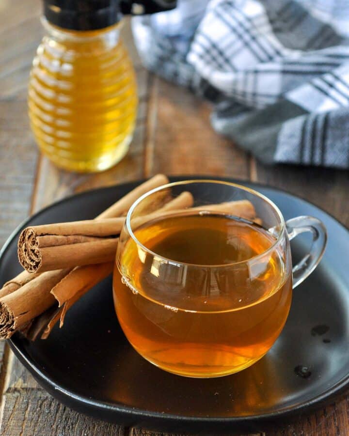 A clear glass mug of cinnamon tea on a black plate with some cinnamon sticks and a honey pot.