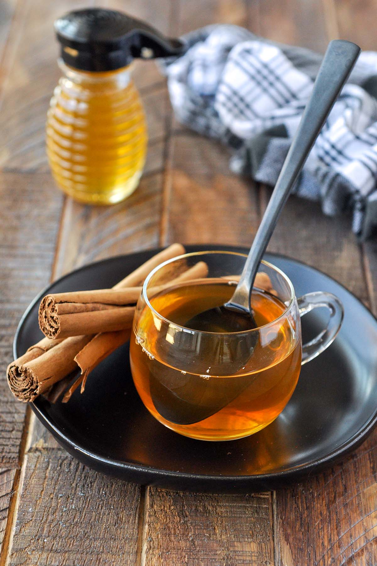 A clear glass mug of cinnamon tea on a black plate with some cinnamon sticks and a honey pot.