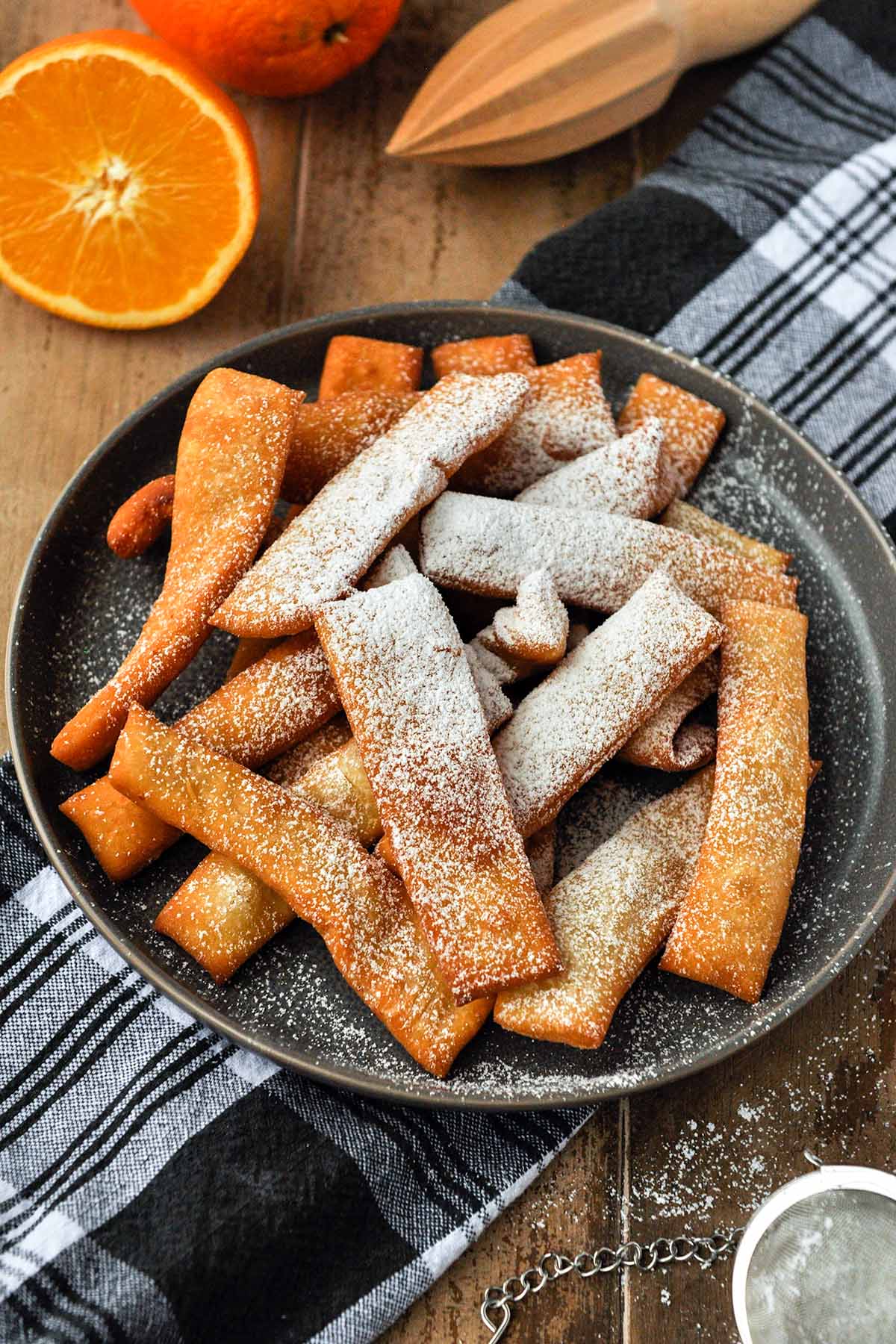 On a black and white towel sits a plate of hojuelas that has been dusted with powdered sugar. Half an orange and a citrus reamer are in the background.
