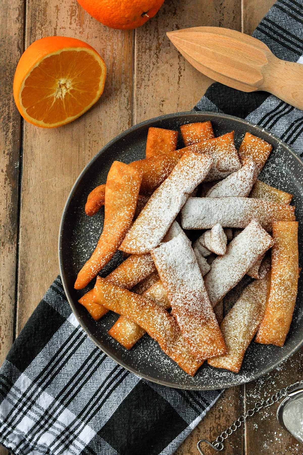 On a black and white towel sits a plate of hojuelas that has been dusted with powdered sugar. Half an orange and a citrus reamer are nearby.