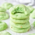 A stack of pistachio cookies, the top one is missing a bite and there are more cookies all around.