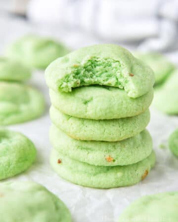 A stack of pistachio cookies, the top one is missing a bite and there are more cookies all around.
