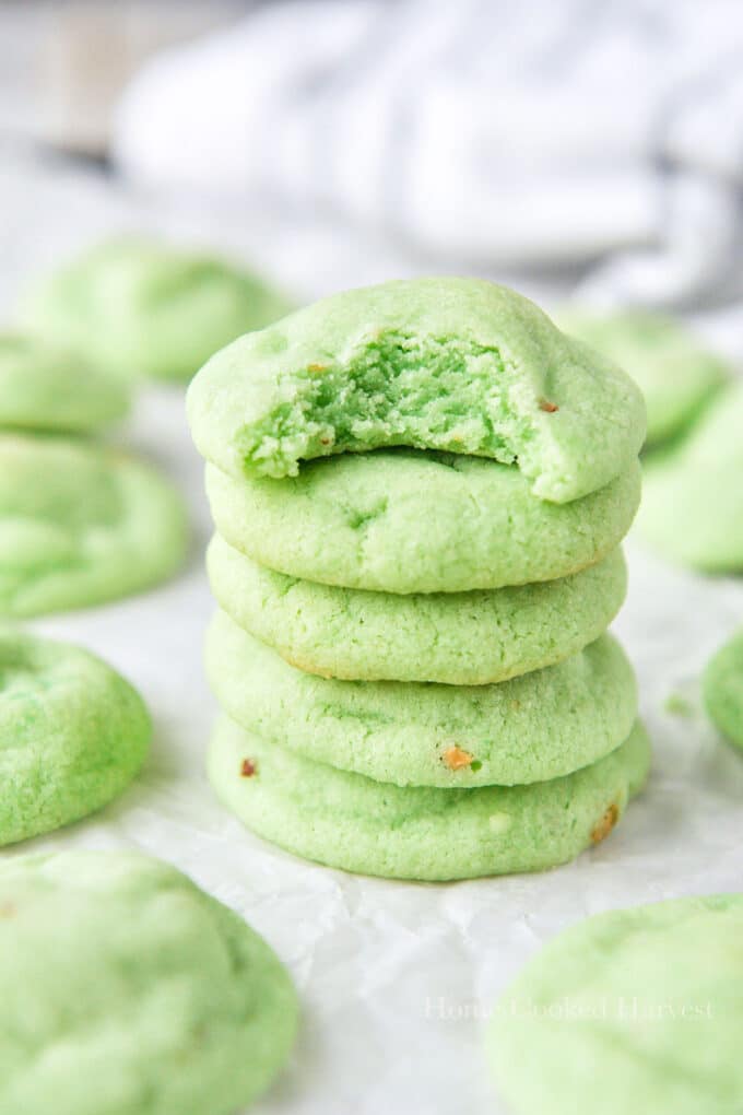 A stack of pistachio cookies, the top one is missing a bite and there are more cookies all around.