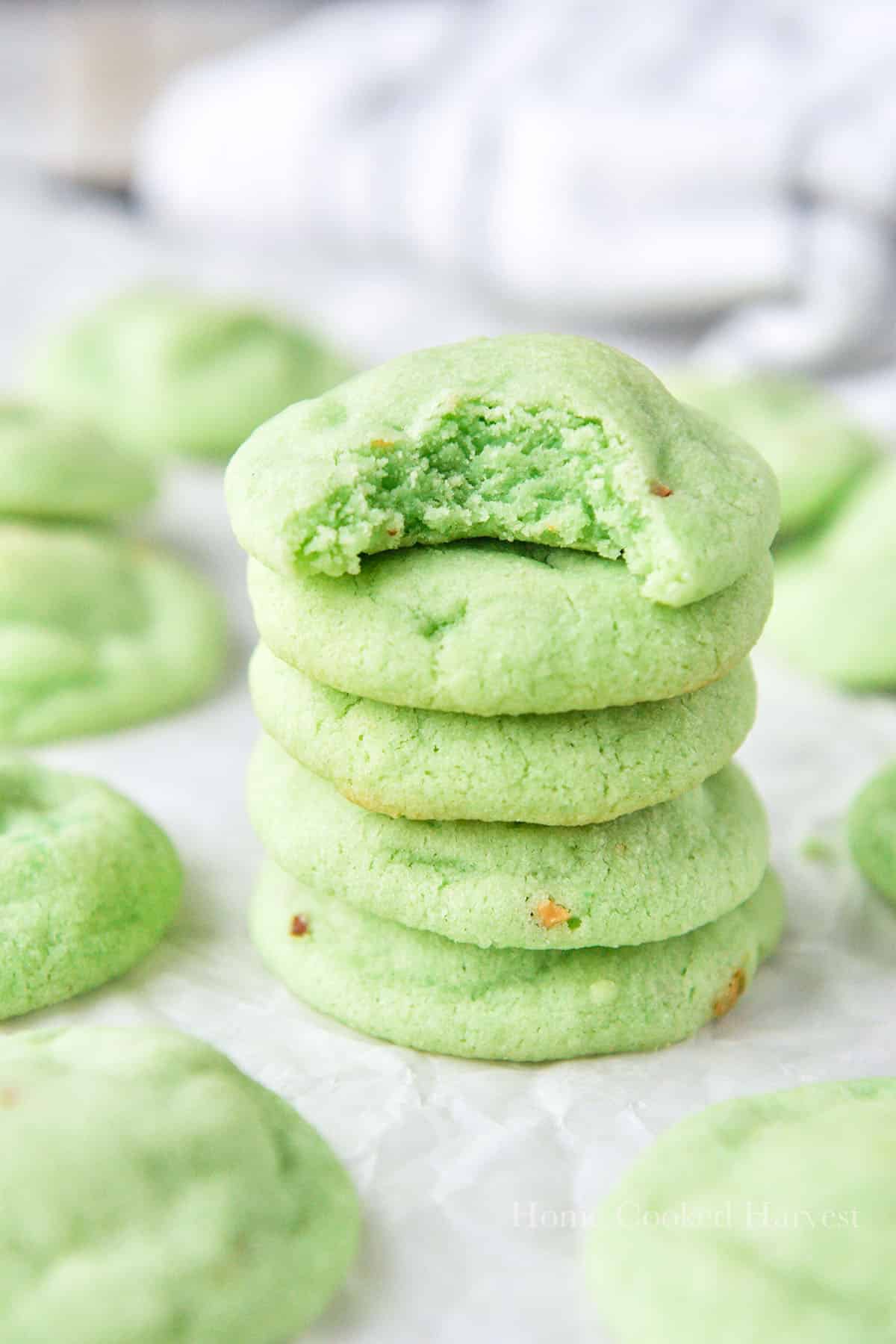 A stack of pistachio cookies, the top one is missing a bite and there are  more cookies all around.