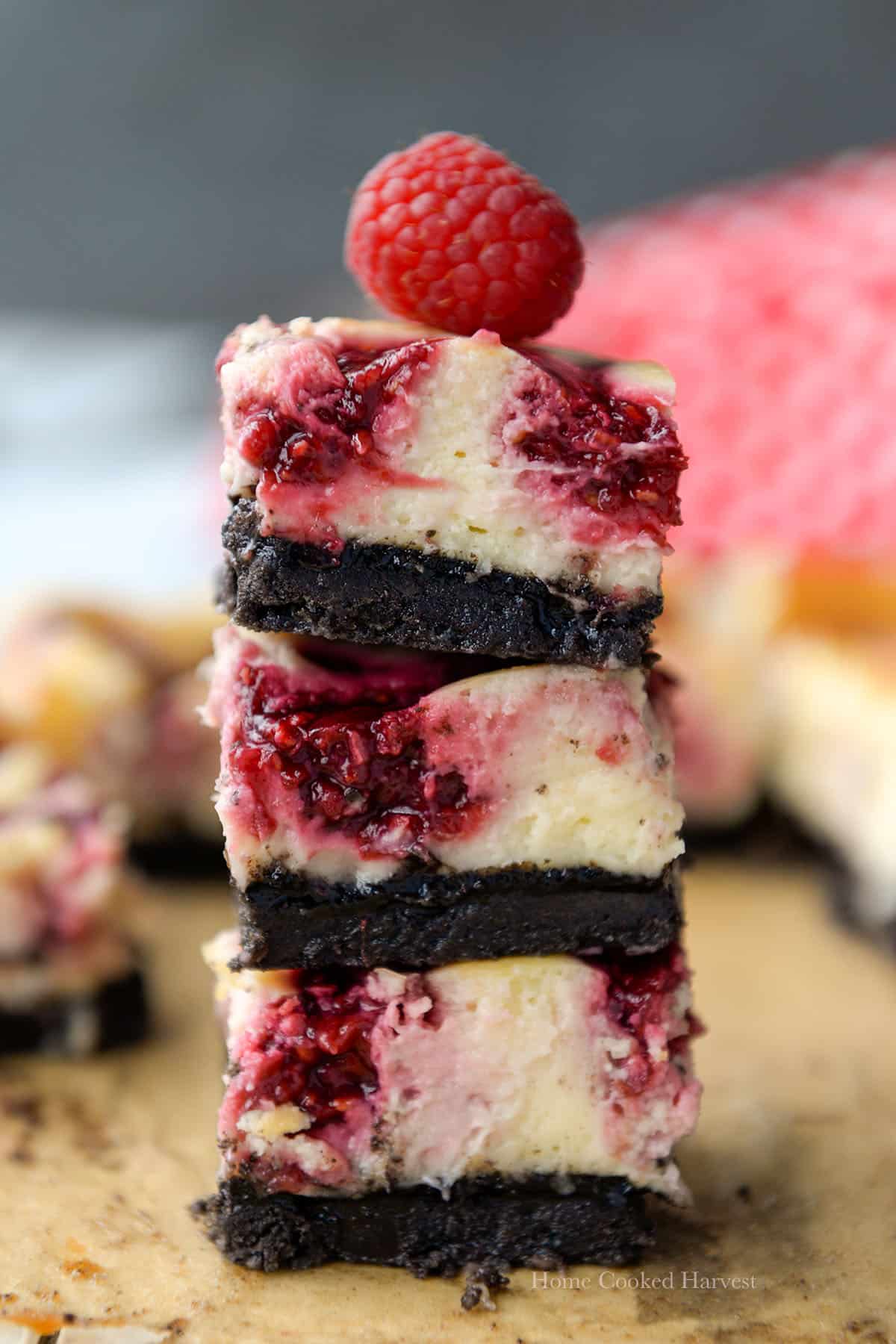 A stack of three raspberry bars with a raspberry on top and a red and white towel in the background.