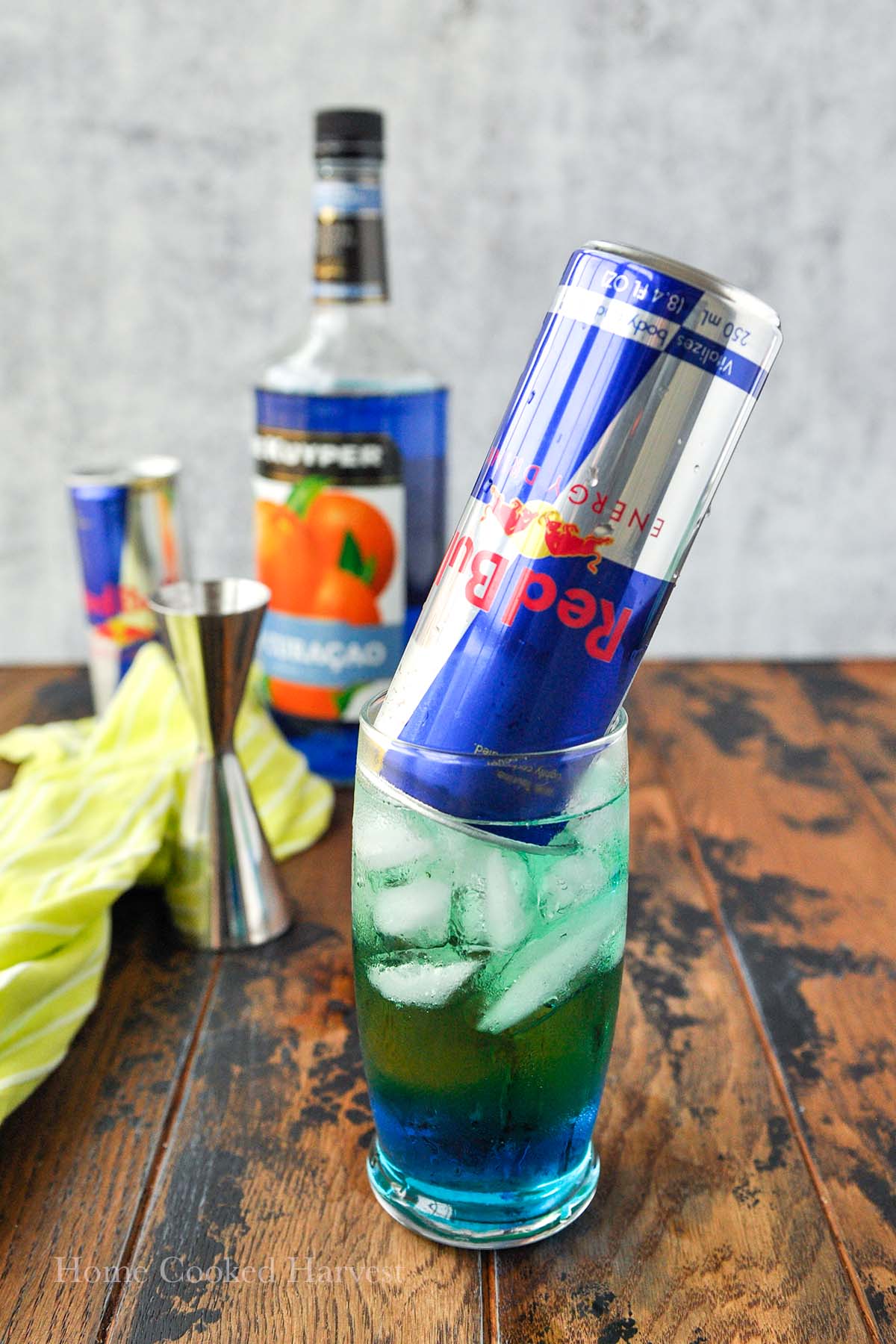 An upside down can of Red Bull in a glass of Irish trash can. Bottle of alcohol, a Red Bull, and jigger in the back ground.