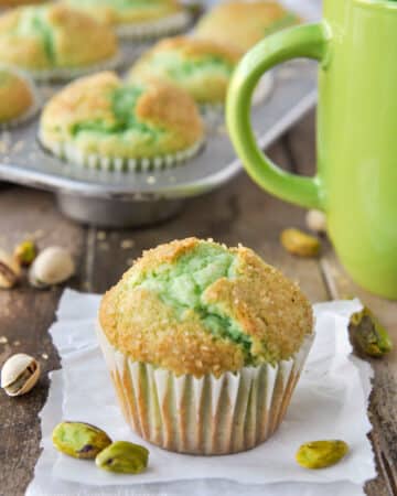 A pistachio muffin on parchment paper, with a green mug and a tray of muffins in the background.