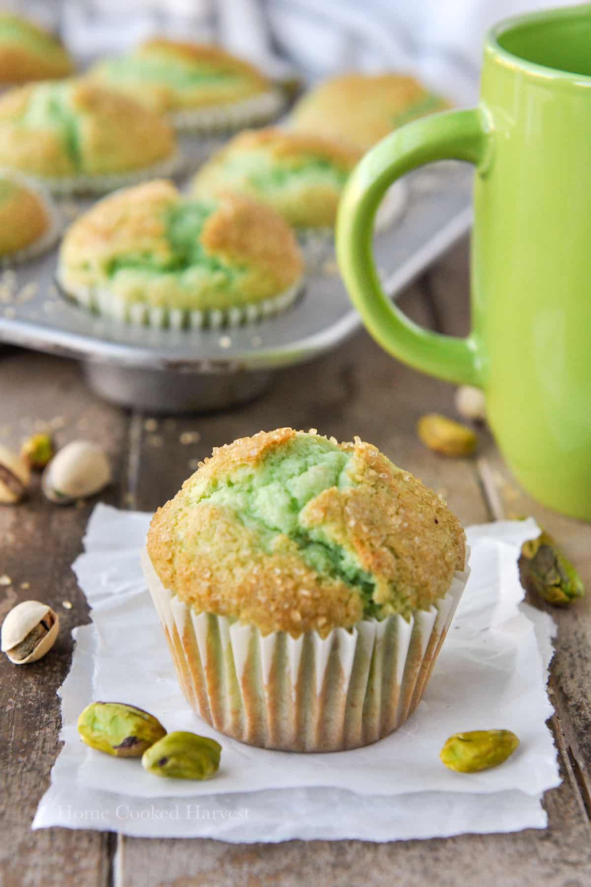 A pistachio muffin on parchment paper, with a green mug and a tray of muffins in the background.