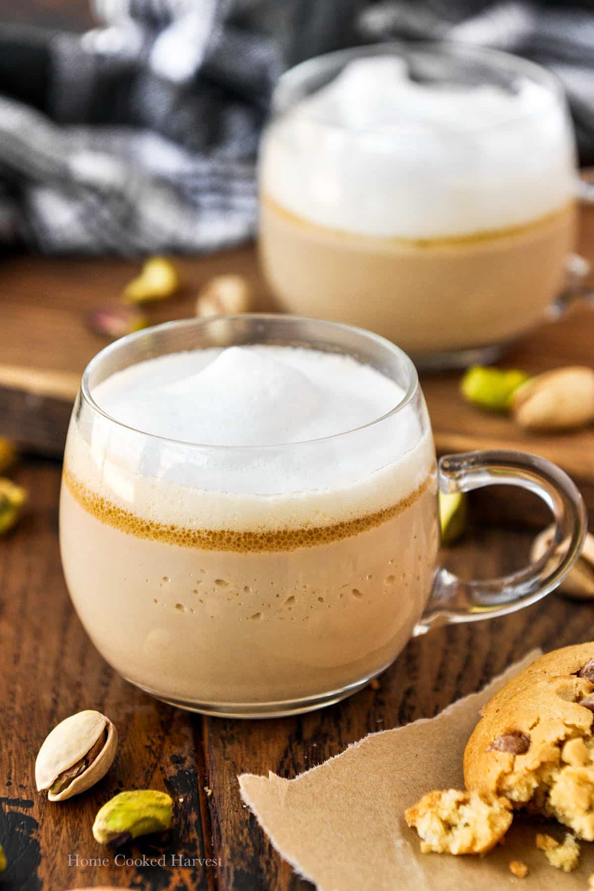 Up close of a mug of pistachio latte with another mug in the background surrounded by fresh pistachios and a cookie.