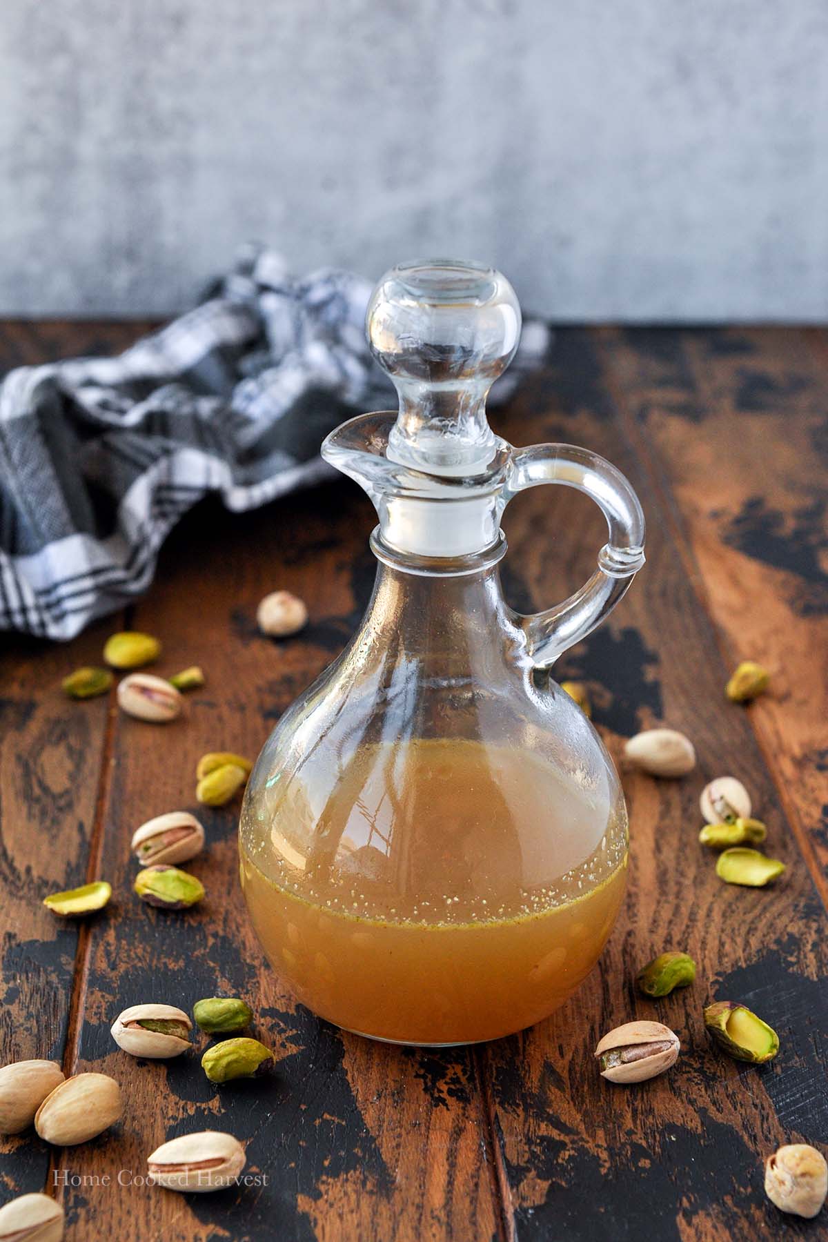 A glass jug filled with pistachio syrup, pistachios around, and a black and white towel.