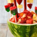 Up close of a watermelon bowl stuffed with chamoy and tajin coated fruits. Lollipops and fruit skewers are stuck into the watermelon rind.