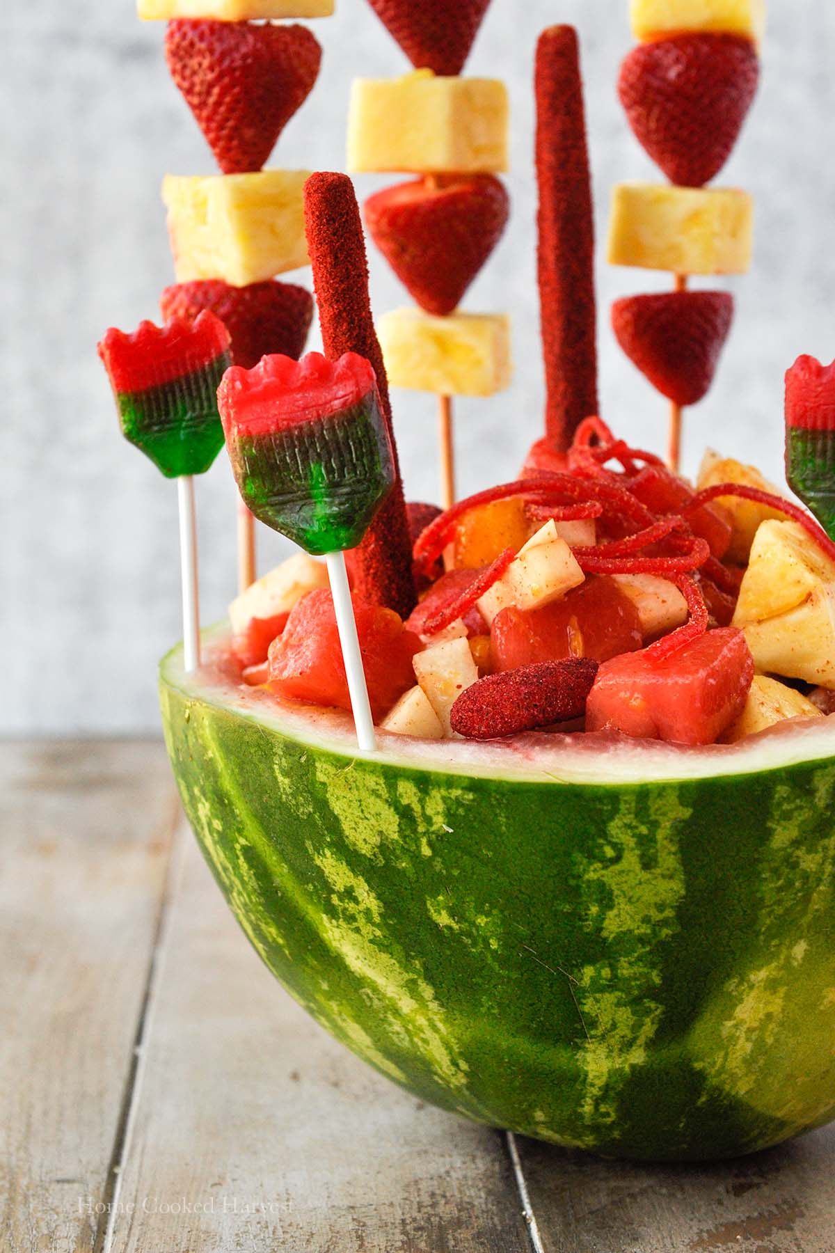 A watermelon bowl stuffed with chamoy and tajin coated fruits. Lollipops and fruit skewers are stuck into the watermelon rind.