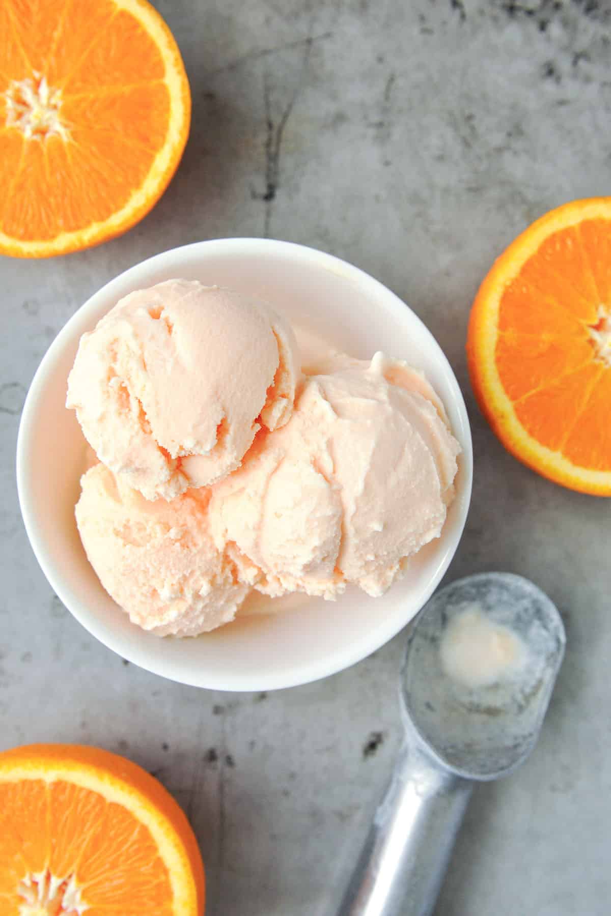Above view of orange ice cream in a bowl with orange halves and an ice cream scoop.
