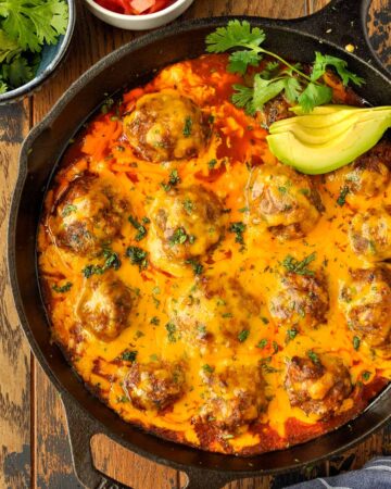 Above view of a skillet of cheesy enchilada meatballs with avocado, cilantro, and tomatoes.