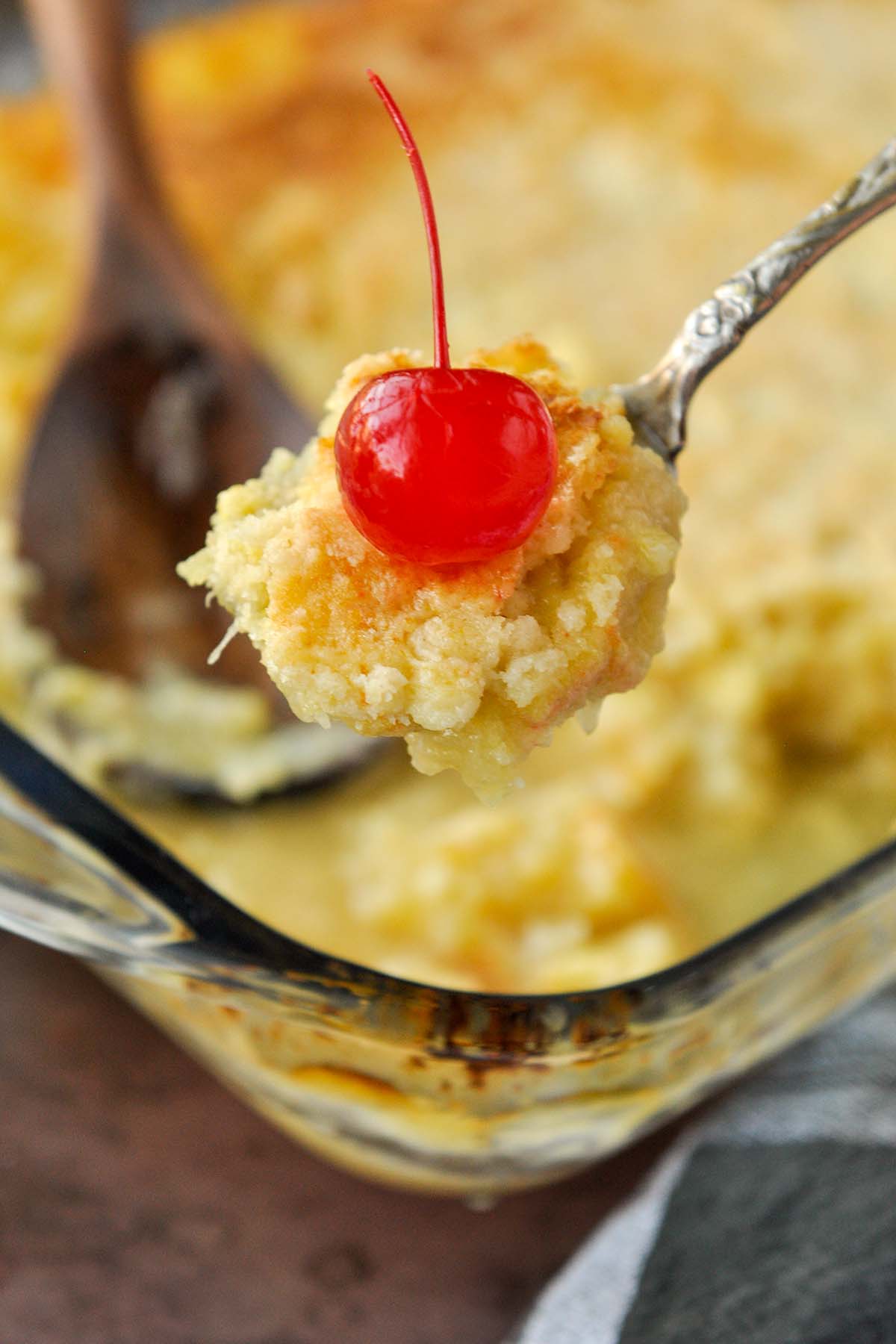 A spoon of dump cake with a cherry over the dish of pineapple dump cake.