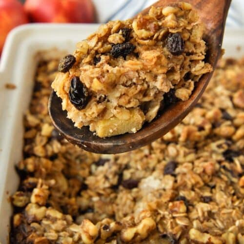 A big spoonful of baked oatmeal over the baking dish with a striped towel and apples in the background.