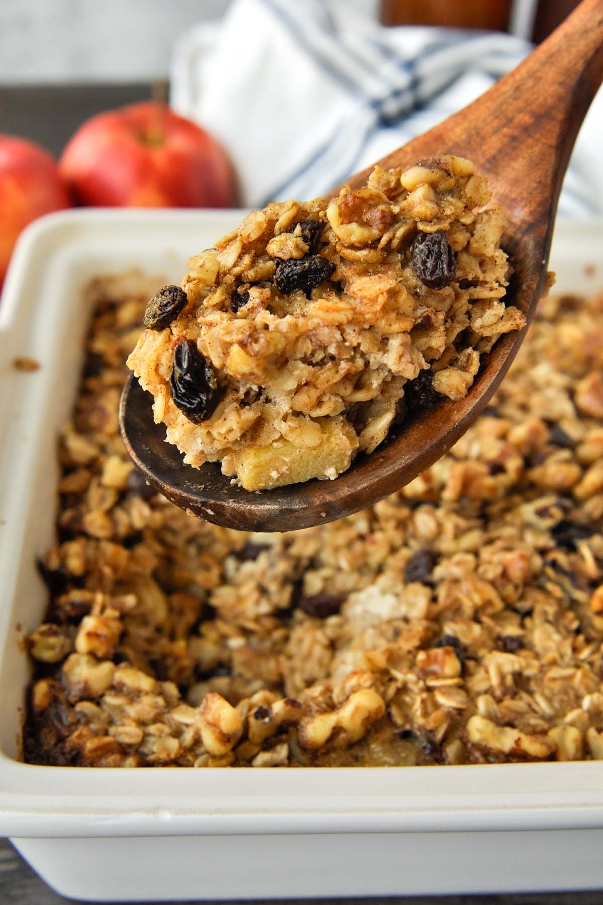 A big spoonful of baked oatmeal over the baking dish with a striped towel and apples in the background.