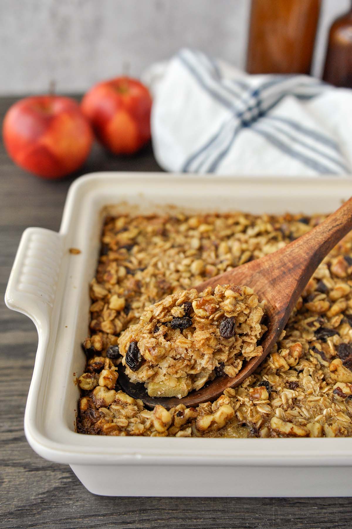 A spoonful of baked oatmeal in a pan of the oatmeal with a towel and apples in the background.
