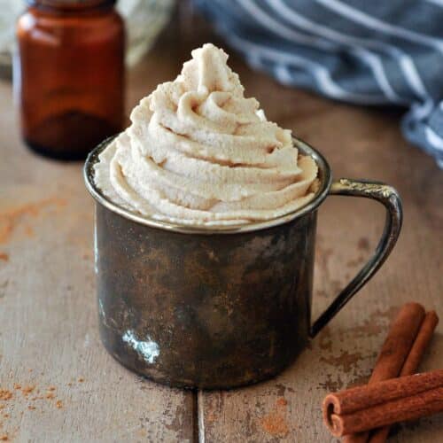A metal cup filled with a swirl of cinnamon whipped cream, glass bottles in the background with a black striped towel and cinnamon sticks up front.