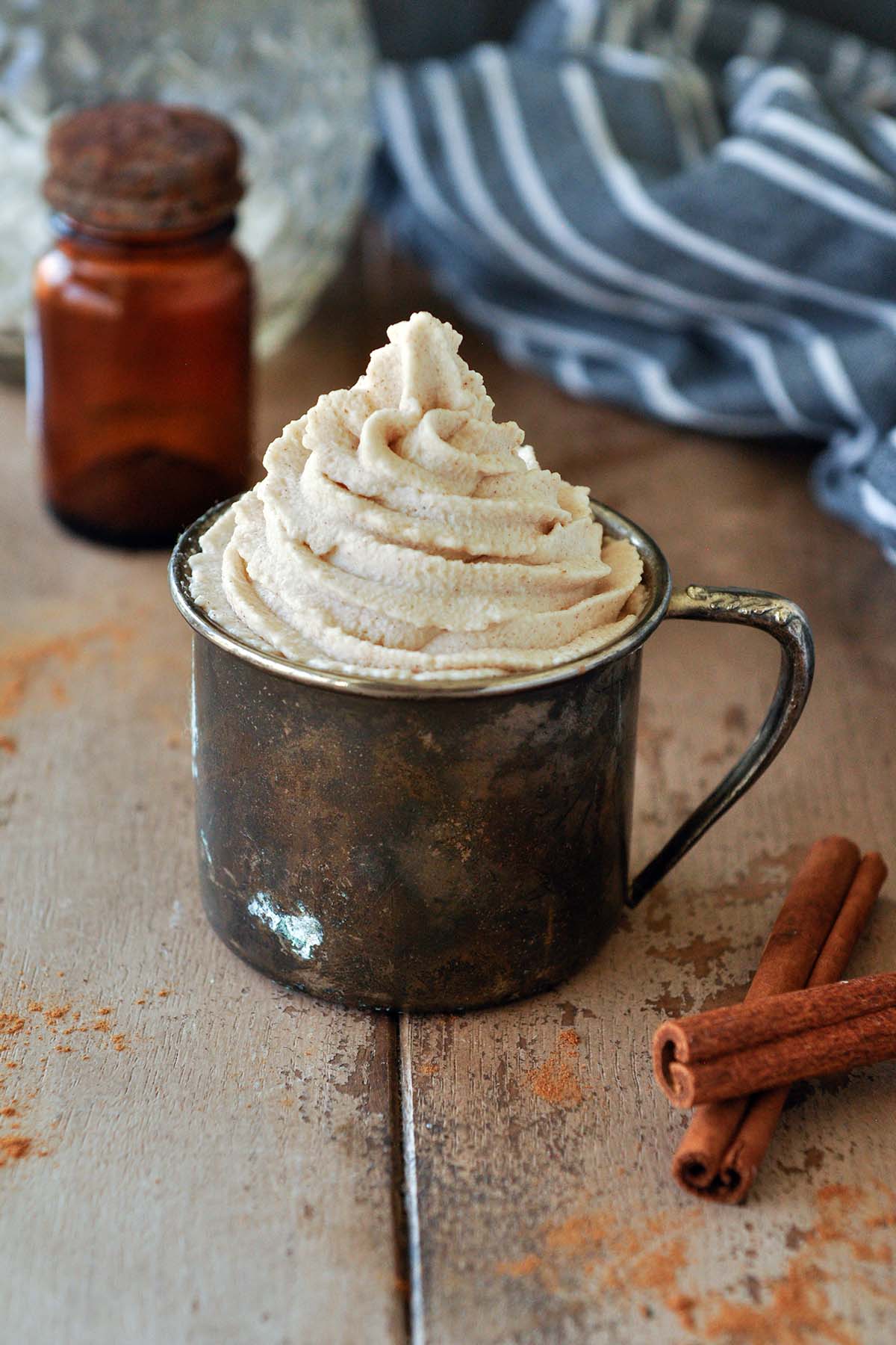 A metal cup filled with a swirl of cinnamon whipped cream, glass bottles in the background with a black striped towel and cinnamon sticks up front.