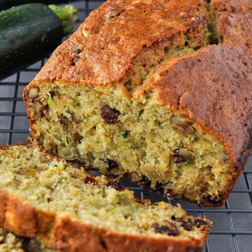 A loaf of pineapple zucchini bread with some slices cut and zucchini in the background.