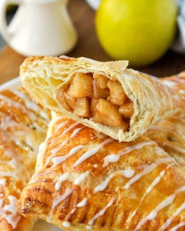 Half of an apple puff pastry on top of other pastries with an apple in the background.