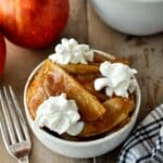 Up close of baked apples in a small white dish with a little whipped cream. Fresh apples and baking dish in the back ground.