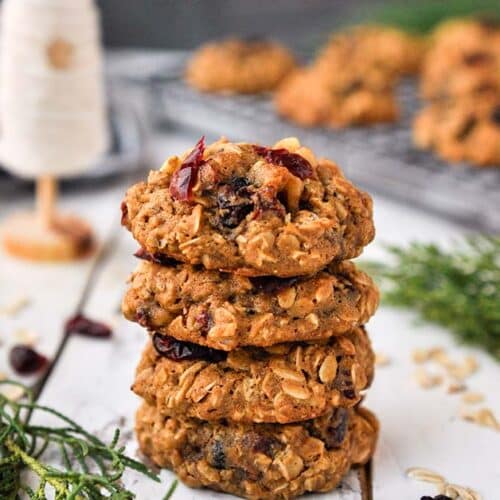 A stack of cranberry walnut oatmeal cookies with a cooling rack of cookies and a small white Christmas tree in the background.