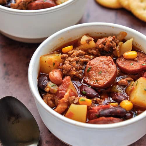 A bowl of cowboy stew with a spoon and some crackers.