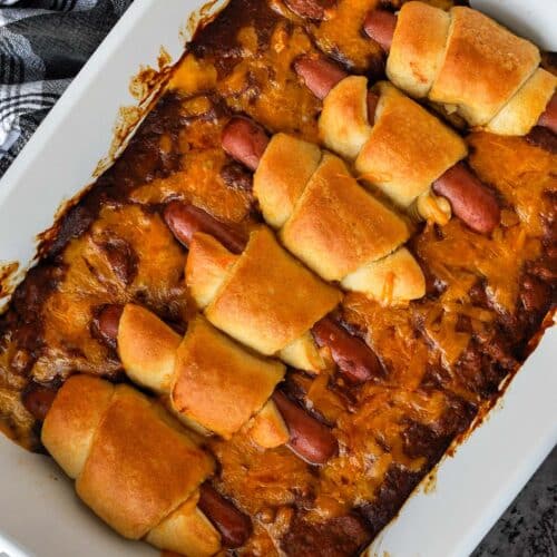 A whole baking dish of hot dog casserole with a small bowl of cheese and a black and white towel.
