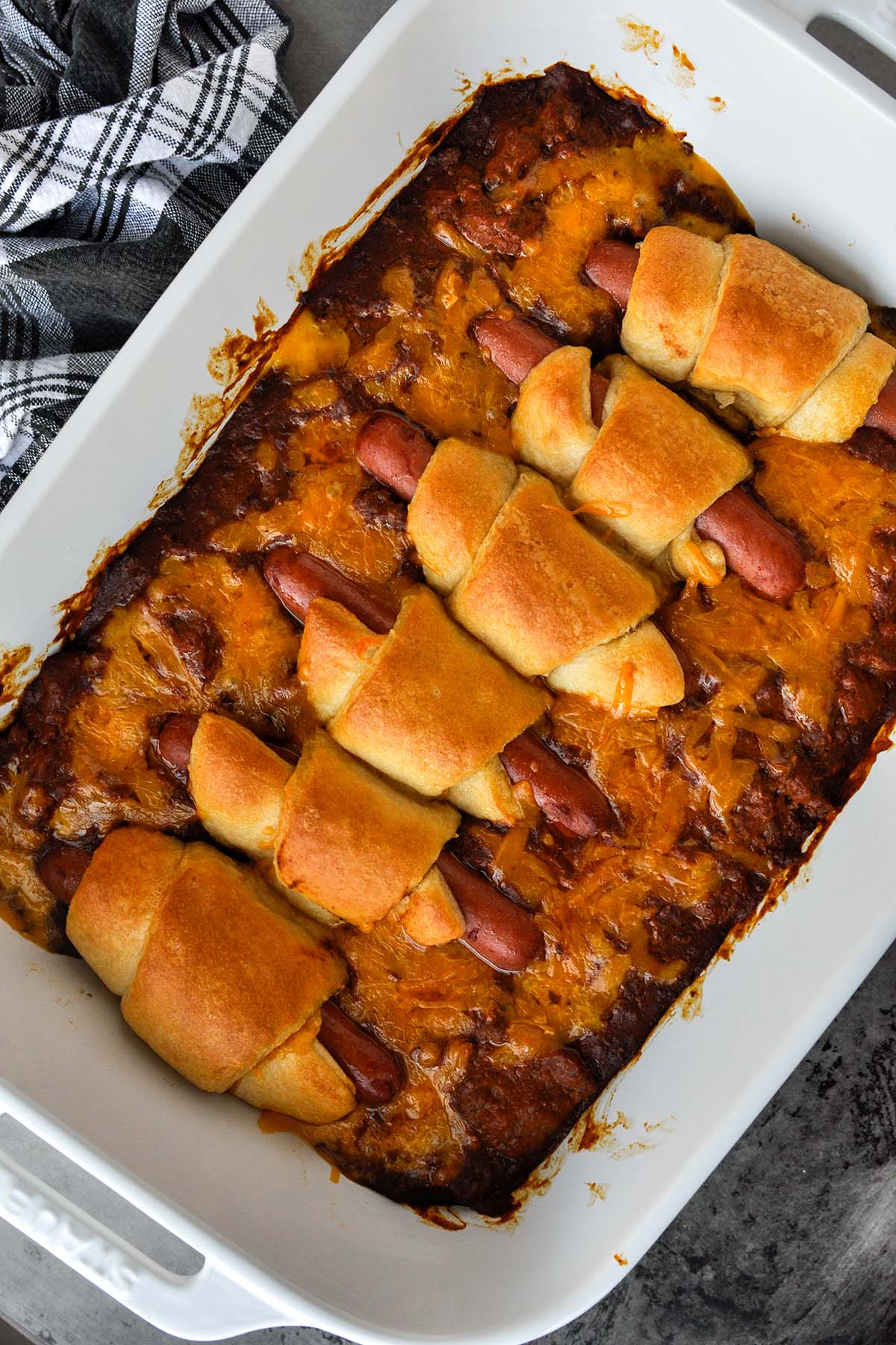 A whole baking dish of hot dog casserole with a small bowl of cheese and a black and white towel.