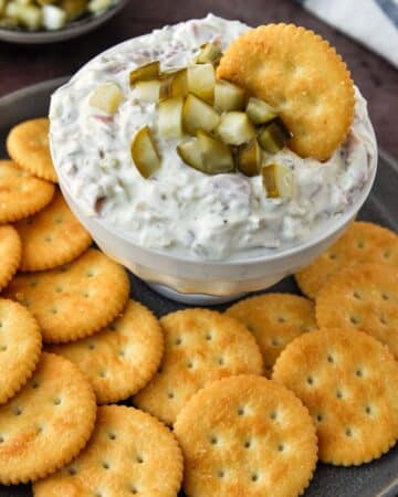A bowl of pickle wrap dip with a cracker on a tray of crackers.