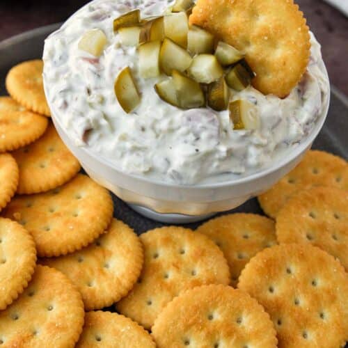 A bowl of pickle wrap dip with a cracker on a tray of crackers.