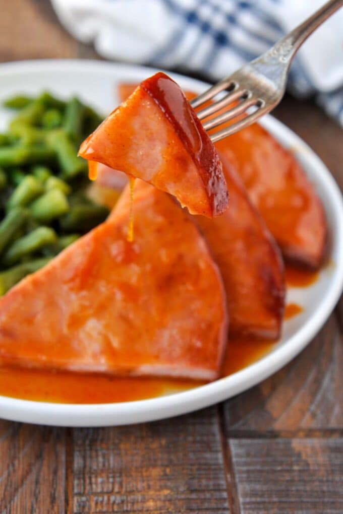 A bite of ham on a fork held above a plateful of ham and green beans.