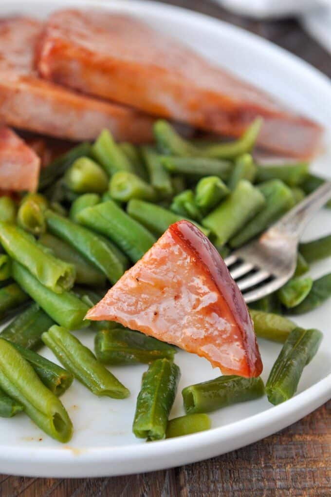 A bite of ham on a fork on a plate of ham slices drizzled with sauce, with a side of green beans.