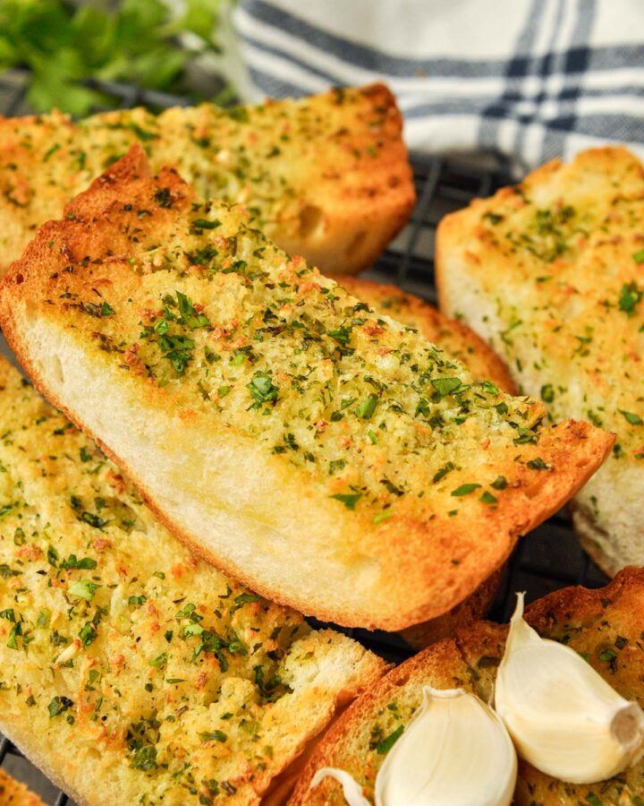 Up close of a chunk of garlic bread surrounded by other pieces of bread and two pieces of garlic. Striped towel in the background.
