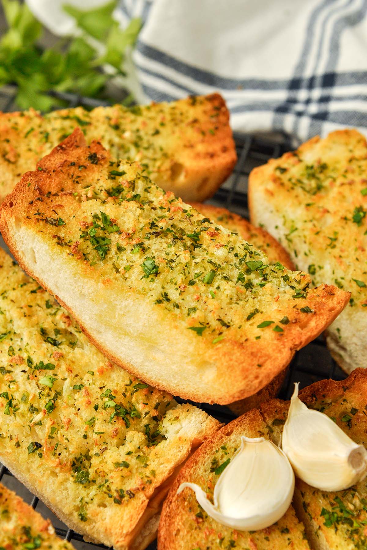 Up close of a chunk of garlic bread surrounded by other pieces of bread  and two pieces of garlic. Striped towel in the background.