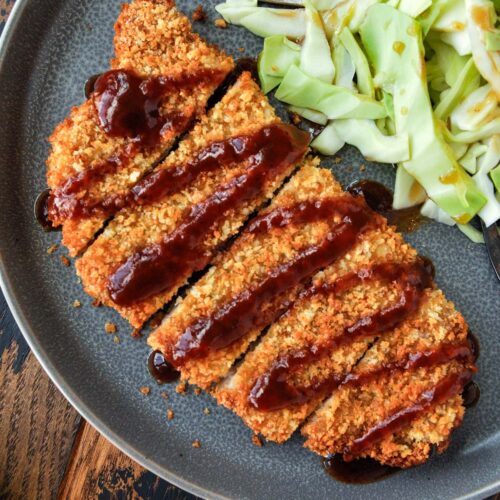 Up close, above view of pork tonkatsu on a plate that has been sliced and has a drizzle of katsu sauce across the top and served with some shredded cabbage.